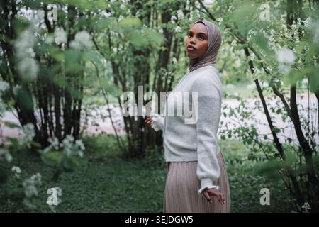 Giovane donna musulmana in hijab tra rami in fiore nel parco primaverile Foto Stock