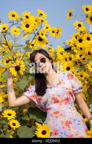 Bella donna asiatica in piedi all'aperto nel campo dei fiori gialli in fiore durante la calda stagione estiva indossando un abito floreale sentendosi rilassata calma e tranquilla Foto Stock