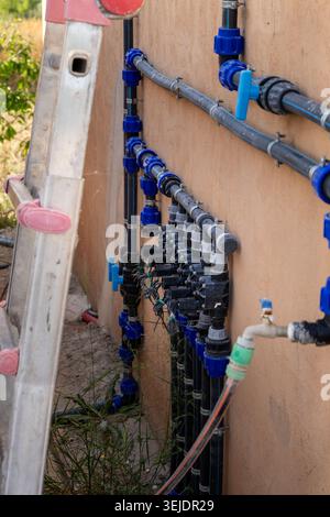 Una vista dettagliata di un sistema di collettore di irrigazione automatizzato con valvole multiple e raccordi per tubi blu montati su una parete esterna, con una scala Foto Stock