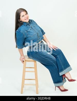 Donna elegante e felice in un outfit in denim che posa su uno sgabello Foto Stock