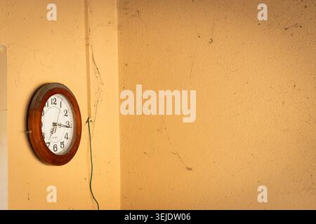Un orologio analogico vecchio stile con una cornice in legno scuro è montato su una parete ruvida color ocra, che mostra segni di età e abbandono Foto Stock