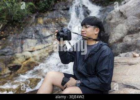 Giovane fotografo asiatico che tiene in mano la macchina fotografica seduto su una roccia bagnata vicino a una cascata che scorre all'aperto, mentre esplora l'avventura con la fotografia naturalistica Foto Stock