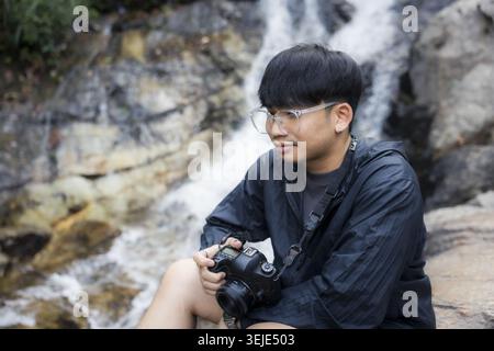 Giovane fotografo asiatico adulto che indossa occhiali trasparenti e giacca nera scura che riposa tenendo la macchina fotografica vicino alla splendida cascata selvaggia nella sensazione della natura Foto Stock