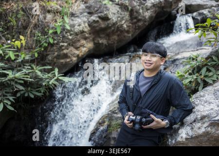 Il giovane fotografo asiatico sorride felicemente tenendo la macchina fotografica in piedi vicino alla splendida cascata naturale ruscello roccioso in un lussureggiante ambiente forestale Foto Stock
