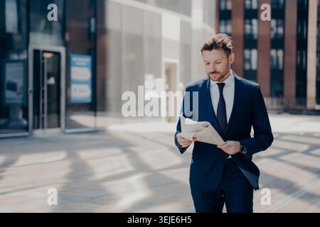 Il giovane uomo d'affari di successo con la barba ordinata in abbigliamento formale legge il giornale mentre cammina per un moderno edificio di vetro nel quartiere finanziario della città nelle soleggiate mattine ventose all'aperto Foto Stock