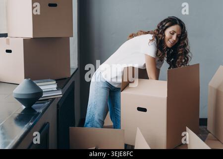 Donna sorridente che confeziona scatole di cartone a casa durante il giorno del trasloco e organizza oggetti personali in luminoso e moderno appartamento interno Foto Stock