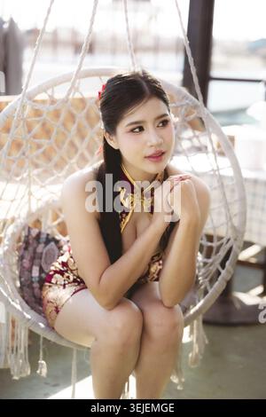 Giovane donna asiatica premurosa in un bellissimo abito rosso cheongsam seduta su una sedia a dondolo sospesa all'aperto, che guarda lontano con espressione pensiva e serena Foto Stock