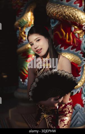 Bella donna asiatica con tradizionale abito cinese qipao rosso e oro, ha un fan nero. Si trova graziosamente vicino alla statua del drago dorato nel tempio, con Foto Stock