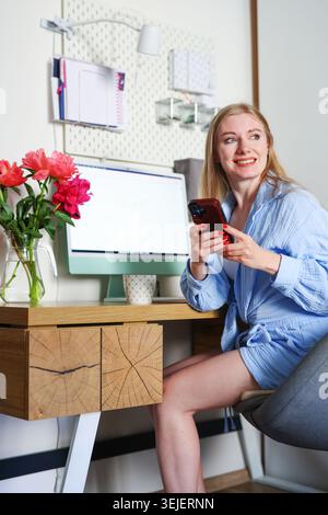 Donna sorridente seduta alla scrivania con smartphone e peonie, moderno ritratto dell'ufficio domestico per la comunicazione e i temi dello stile di vita digitale. Foto Stock