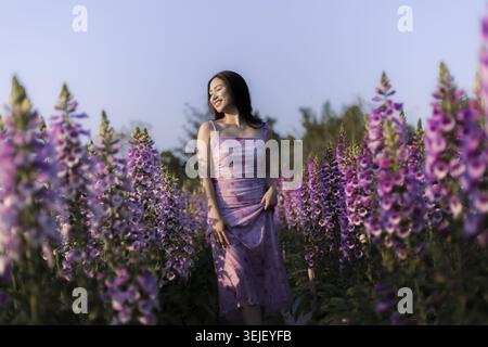 Donna felice sorridente in abito viola in piedi in un campo di fiori all'aperto. Ama la natura estiva, circondata da splendidi fiori viola durante il seren Foto Stock