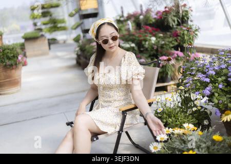 Una giovane donna asiatica serena, con un bel vestito alla moda e un berretto, si siede sulla sedia della serra, godendosi un'atmosfera tranquilla e toccando i fiori in giardino Foto Stock