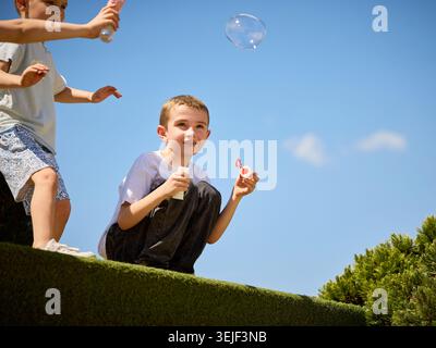 Ragazzo che soffia bolle di sapone sotto il cielo azzurro limpido nel parco estivo. Foto Stock