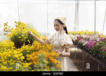 Elegante donna asiatica che gode di un momento sereno tendente al fiore giallo in serra. giovane giardiniere che si prende cura delle piante colorate in un bellissimo vivaio Foto Stock
