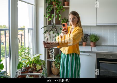 Influencer femminile che registra un blog a tema sull'impianto al telefono con Alocasia bambino in mano, in live streaming Foto Stock