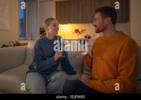 Sorridente coppia felice brindando con bicchieri da vino, rilassandosi sul divano in una serata romantica a casa Foto Stock