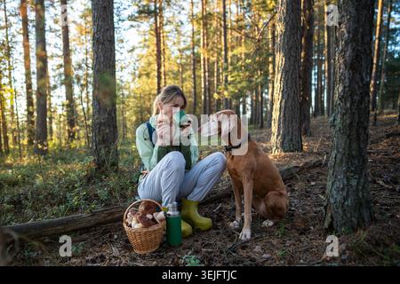 Donna serena che beve tè caldo nella foresta, mentre il fedele cane Vizsla guarda attentamente nelle vicinanze implorare il cibo Foto Stock