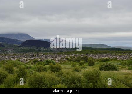 Una vista lontana del paesaggio di un cratere nero a cono vulcanico e montagne sotto basse nuvole ricoperte attraverso verdi pianure arbustive in Islanda. Foto Stock