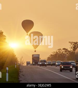 Ripresa verticale di due mongolfiere in silhouette che salgono su un'autostrada con traffico mattutino. La luce calda e nebulosa suggerisce l'alba o il primo giorno. Foto Stock