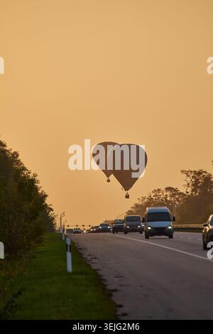 Ripresa verticale di due mongolfiere in silhouette che salgono su un'autostrada con traffico mattutino. La luce calda e nebulosa suggerisce l'alba o il primo giorno. Foto Stock