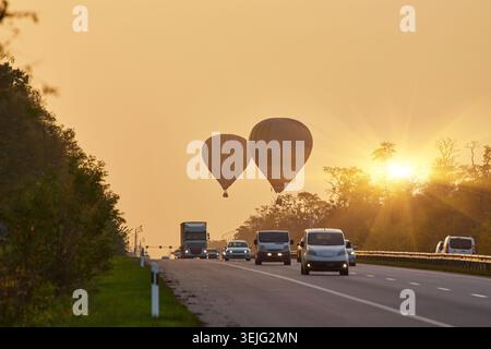Due mongolfiere si stagliano contro un cielo fosco all'alba o al tramonto, fluttuando sopra un'autostrada trafficata con auto e camion. Gli alberi fiancheggiano la strada. Foto Stock