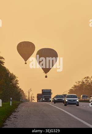 Ripresa verticale di due mongolfiere in silhouette che salgono su un'autostrada con traffico mattutino. La luce calda e nebulosa suggerisce l'alba o il primo giorno. Foto Stock