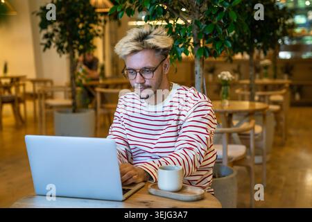 Un uomo concentrato in remoto lavora con un computer portatile al tavolo del bar. Libero professionista che lavora online in remoto Foto Stock