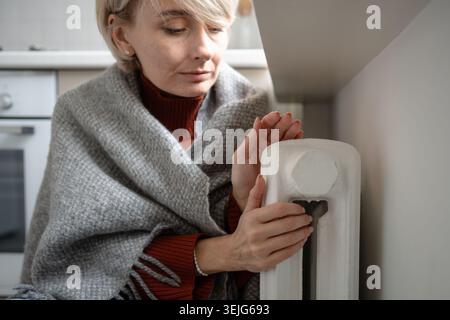 Stanca donna di mezza età che si gela in plaid e che riscalda le mani sul radiatore del riscaldamento centrale nell'appartamento Foto Stock