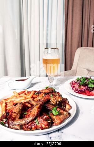 Deliziose ali di pollo alla griglia, con patatine fritte, insalata e una bevanda rinfrescante, a un tavolo accogliente, non ci sono persone Foto Stock