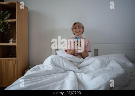 Allegra donna di mezza età seduta a letto e procrastinare la mattina scorrendo i social media. Foto Stock
