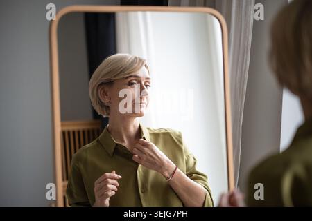 Una donna bionda di mezza età che si prepara per l'appuntamento, controlla la prospettiva davanti allo specchio a casa. Foto Stock