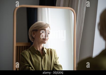 Bella donna bionda di mezza età in piedi davanti allo specchio sorridente e si gode il proprio riflesso. Foto Stock