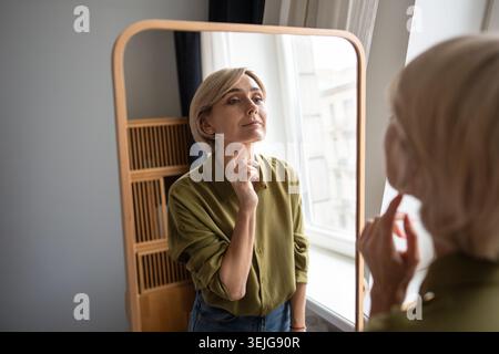 Sorridente bella donna di mezza età in piedi a controllare il trucco allo specchio con un'espressione positiva. Foto Stock