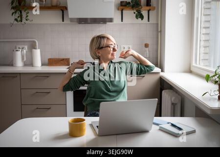Donna d'affari felice di mezza età che riposa su una sedia che si allunga, mentre lavora a distanza da casa. Foto Stock