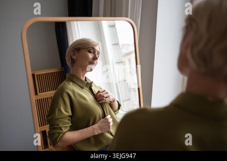 Donna di mezza età in piedi davanti allo specchio provando nuovi vestiti, controllando la prospettiva nella riflessione. Foto Stock