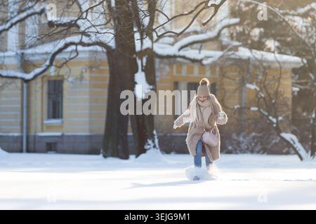 Bella e gioiosa giovane donna con cappotto beige, cappello con pompon che cammina, divertendosi in un parco innevato Foto Stock