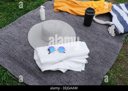 Accessori da spiaggia con borsa da spiaggia, cappello, occhiali da sole e bottiglia di crema solare sulla coperta sull'erba verde Foto Stock