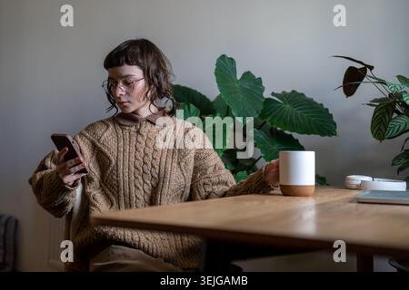 Studentessa teenager concentrata e penosa seduta a tavola da sola con la tazza, leggendo notizie online, scorrendo foto, social network in Internet, messaggistica Foto Stock