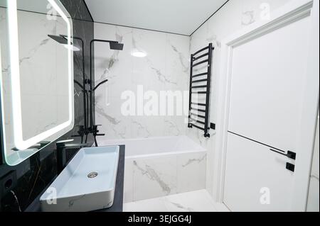 Bagno moderno con lavabo rettangolare, vasca con doccia a parete. Piastrelle di marmo bianco, accenti neri creano interni puliti e minimali, concetto di rinnovamento contemporaneo, soggiorno confortevole Foto Stock