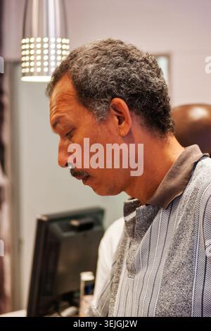 la vista laterale del profilo mostra un uomo maturo di corse miste con baffi e capelli di sale e pepe in attesa pazientemente in coda presso un banco di assistenza o un banco di vendita Foto Stock