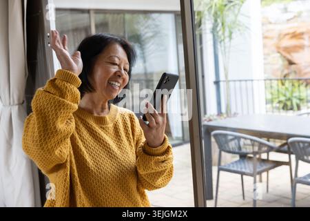 Donna asiatica anziana vicino a una porta di vetro scorrevole in casa sorridente, sventolando allo smartphone con mobili da patio. Senior, allegro, digitale, Foto Stock