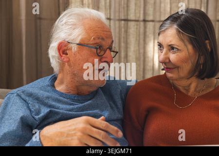 Una coppia anziana seduta sul divano a casa, che parla e sorride, indossa occhiali da vista, maglieria, collana. Pensione, compagnia, c Foto Stock