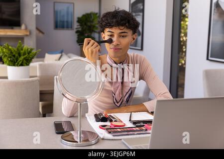 Uomo che applica il trucco con una spazzola al tavolo da pranzo in una casa moderna con specchio, tavolozze, computer portatile. Bellezza, cura del corpo, cosmetici, stile di vita, eleganza, interio Foto Stock