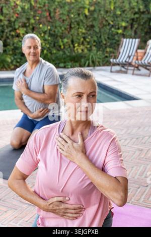 Coppie senior che praticano il traspiramento su tappetini da yoga accanto alla piscina residenziale, indossando abbigliamento sportivo informale. Meditazione, respiro, benessere Foto Stock
