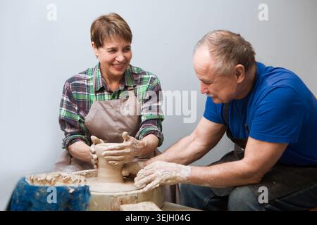 Coppia senior in abiti informali e grembiuli che preparano pentole di ceramica su ruota di ceramica in officina. hobby sulla pensione Foto Stock