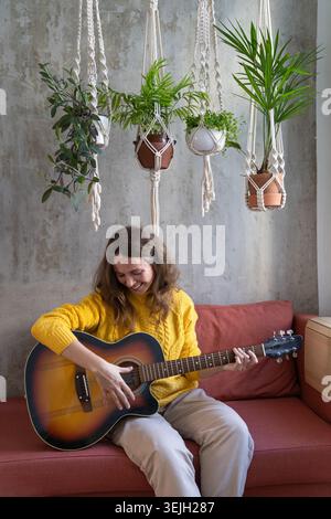 Donna sorridente freelance in maglione giallo, riposante, seduta sul divano sotto un grappolo di piante di cotone macrame con piante d'appartamento, gioca Foto Stock