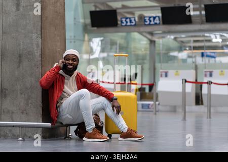 Sorridente giovane hipster afroamericano con valigia che chiama al telefono in attesa di volo in aeroporto. Foto Stock