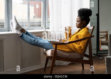 Donna afroamericana freelance seduta su una sedia, gambe appoggiate sul davanzale, studio a distanza, lavoro online su un laptop, divertimento Foto Stock