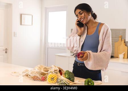Donna dell'Asia meridionale che parla su smartphone, tiene la pesca rossa in cucina con borsa a rete, spazio copia. Moderno, stile di vita, minimalismo, benessere, domestico, na Foto Stock