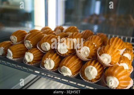 File di pasticcini Choux freschi ripieni di panna montata su uno scaffale espositivo all'interno di una panetteria o pasticceria. Foto Stock