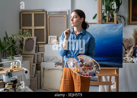 Artista donna pensiva che dipinge foto in casa galleria di workshop, pensando e traendo ispirazione. Foto Stock
